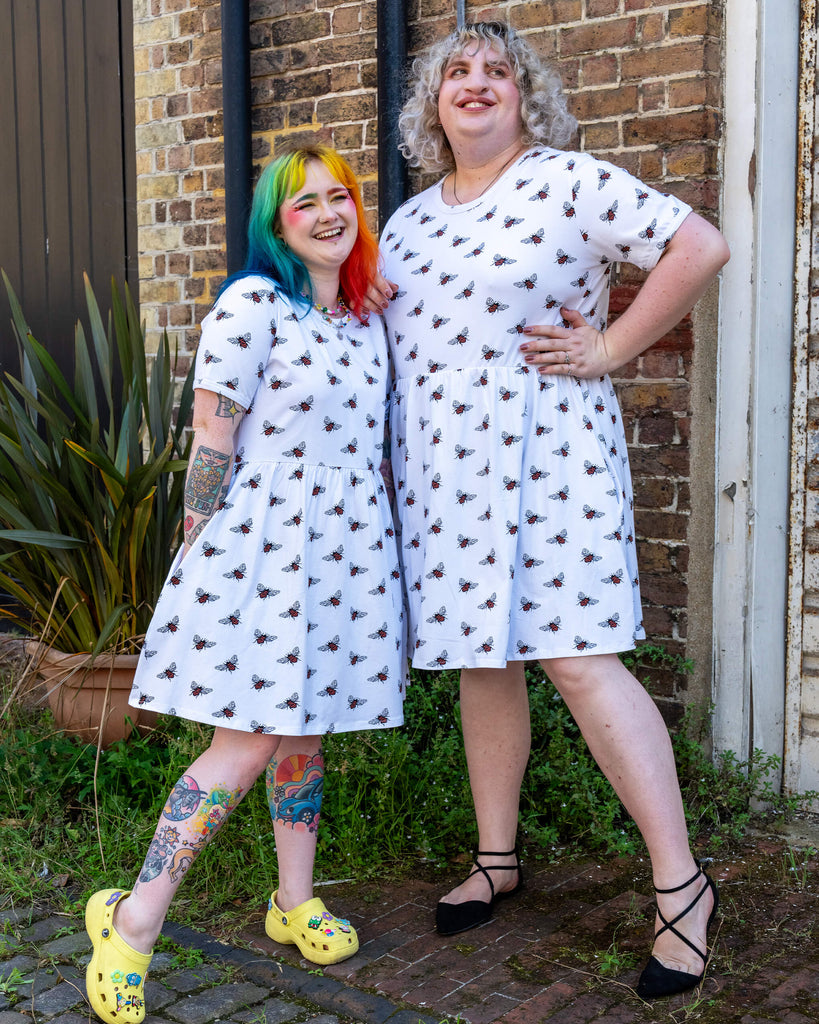 Two friends laughing and posing wearing white oversized smock dresses with an all over bee print from slow, ethical fashion brand Run & Fly. The individual on the left sports rainbow-coloured hair and yellow Crocs, and the individual on the right has curly blonde hair and black shoes.