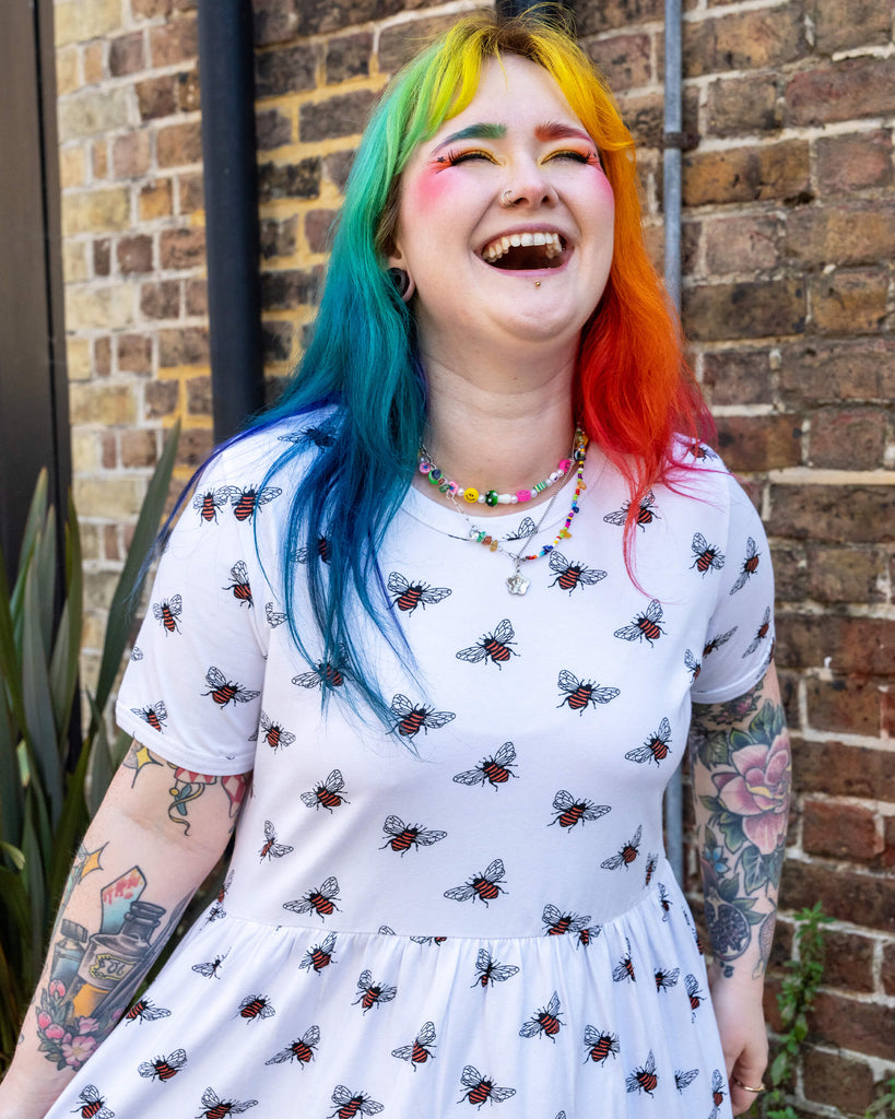 A model with rainbow-coloured hair and bright makeup, wearing a white oversized smock dress with an all over bee print from slow, ethical fashion brand Run & Fly. They are smiling joyfully, with tattoos on their arms looking off to one side.