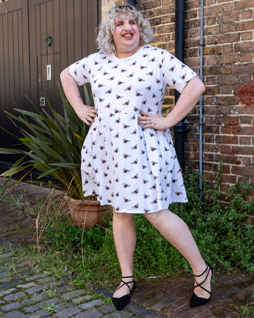 A person with curly blonde hair posing with their hands on their hips, wearing white oversized smock dress with an all over bee print from slow, ethical fashion brand Run & Fly. They are standing in front of a brick wall, smiling confidently.