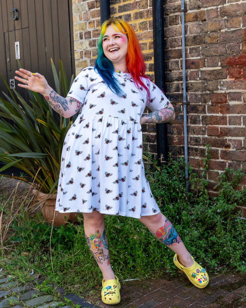 A person with rainbow-coloured hair and bright makeup posing outdoors laughing with their arms out by their sides and one leg slightly bent. They are wearing a white oversized smock dresses with an all over bee print from slow, ethical fashion brand Run & Fly paired with yellow Crocs.