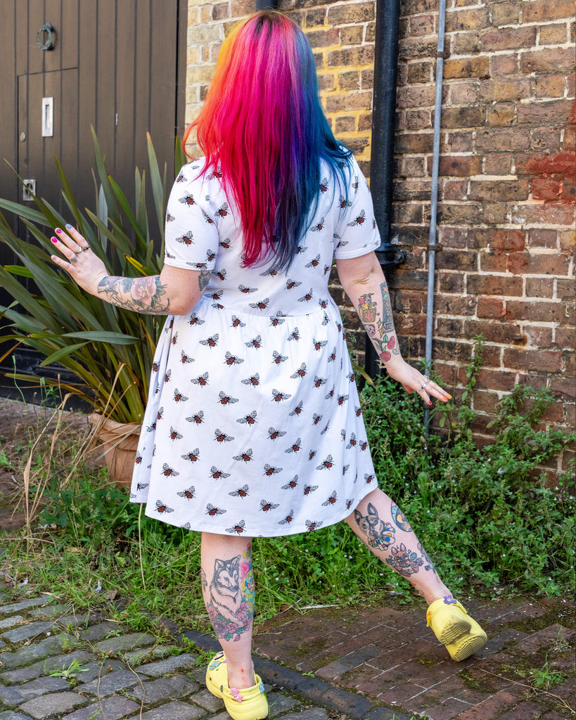 A person with rainbow-coloured hair standing with their back to the camera, showing off the white oversized smock dress with an all over bee print from slow, ethical fashion brand Run & Fly. The model has colourful leg tattoos.