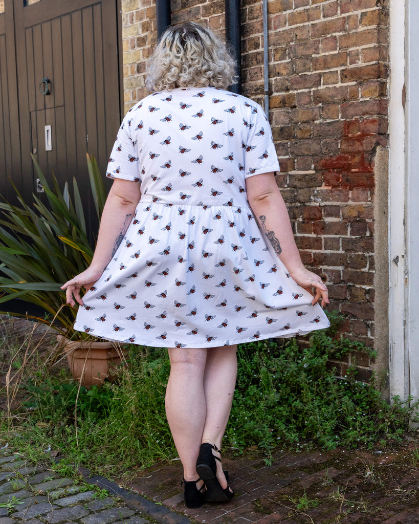 A person with curly blonde hair facing away from the camera, showcasing the back of white oversized smock dress with an all over bee print from slow, ethical fashion brand Run & Fly. They are holding the sides of the dress out slightly, standing in front of a brick wall and plants, wearing black shoes.