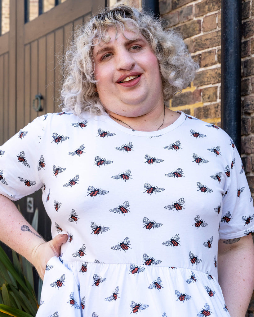 A close-up of a model with curly blonde hair posing with their hand on their hip and other hand in their hair, wearing a white oversized smock dresses with an all over bee print from slow, ethical fashion brand Run & Fly. They are standing against a wooden backdrop.