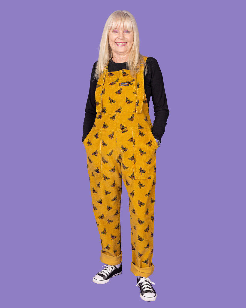 A full-body front view of an elderly woman with long blonde hair wearing a mustard yellow corduroy dungaree with black bee patterns over a black long-sleeve shirt. She has her hands in the front pockets of the dungarees and is wearing black and white sneakers. The background is a solid purple.