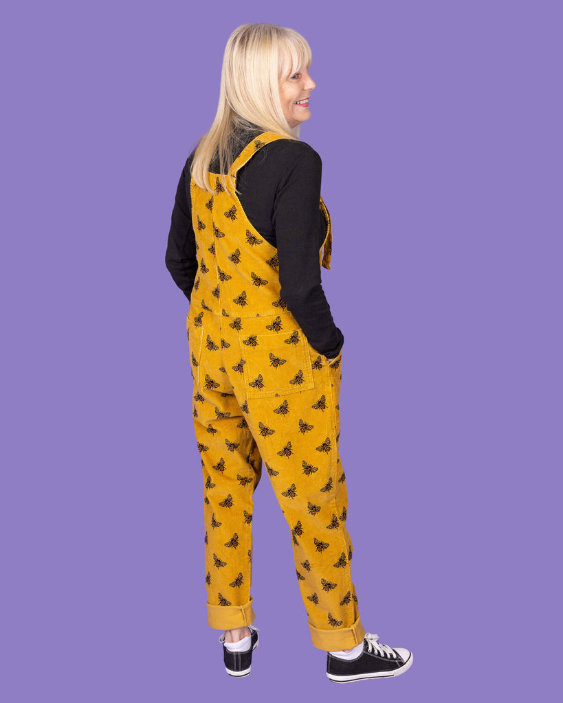 The back view of an elderly woman with long blonde hair wearing a mustard yellow corduroy dungaree with black bee patterns over a black long-sleeve shirt. She has her hands in the back pockets of the dungarees and is standing with one foot slightly forward. The background is a solid purple.