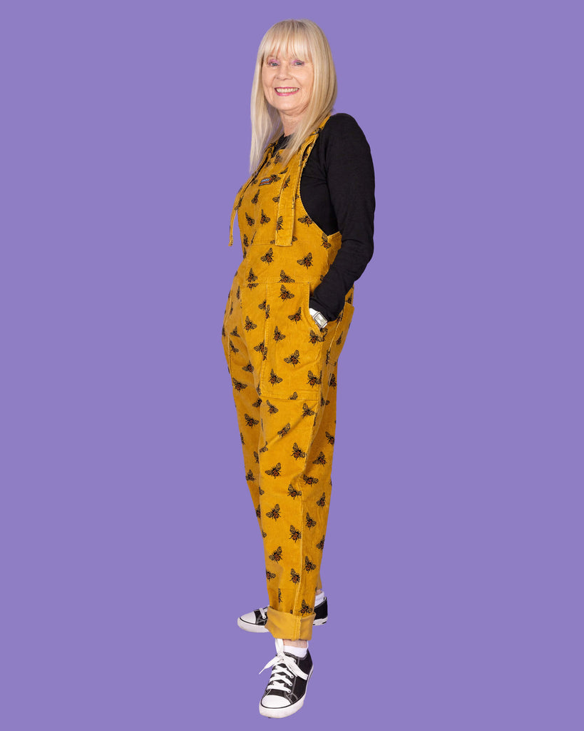 A full-body side profile of an elderly woman with long blonde hair wearing a mustard yellow corduroy dungaree with black bee patterns over a black long-sleeve shirt. She has her hands in the pockets of the dungarees and is wearing black and white sneakers. The background is a solid purple.