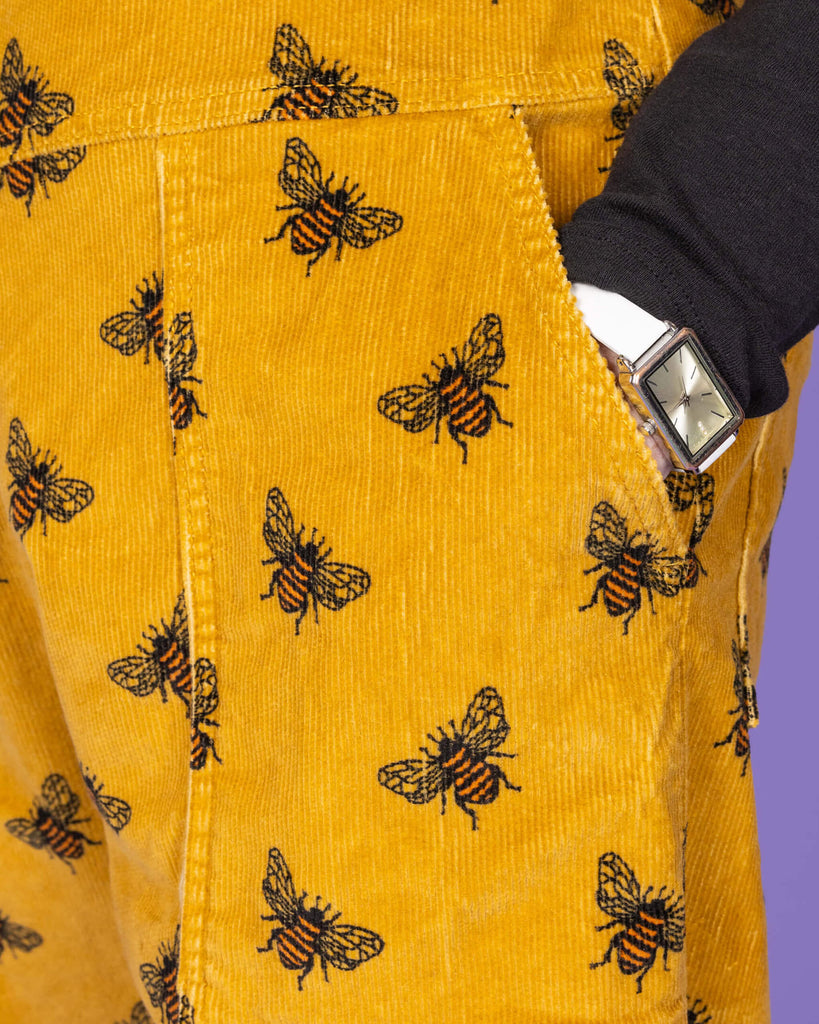A close-up shot of the mustard yellow corduroy dungaree with black bee patterns. The focus is on the pocket and the side, showing a silver wristwatch on the person’s left wrist and the texture of the fabric. The background is a solid purple.