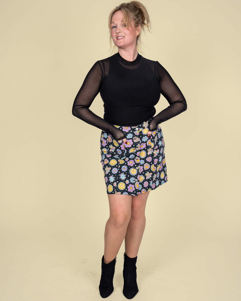 Karina models the Run & Fly mini skirt featuring a vibrant print by Waffle Mama Design. The black skirt is adorned with cheerful smiley faces, colourful flowers, hearts, and playful bees, paired with a sleek black mesh top.