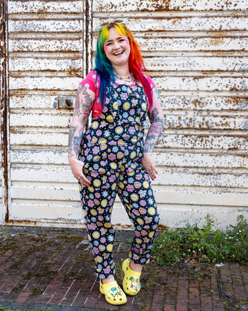 Person with rainbow hair wearing Run & Fly dungarees with a colourful smiley face and flower pattern, paired with a pink polka dot top and yellow Crocs. They pose confidently against a rusted metal wall, showcasing tattoos and vibrant makeup. Run & Fly is a small ethical slow fashion brand known for its quirky, bold prints and creative, alternative style.