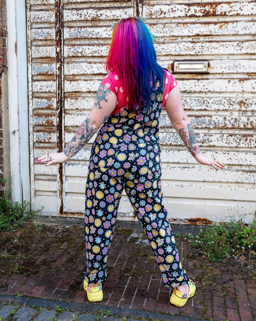 Back view of a person with vibrant rainbow hair, wearing black dungarees with a playful pattern of smiley faces, flowers, and hearts, paired with a red polka dot top. The dungarees are from Run & Fly, a small ethical slow fashion brand known for their fun, colourful prints. The person stands with arms outstretched, showing off their tattooed arms and bright yellow platform shoes. The setting is against a rusted, textured metal wall.