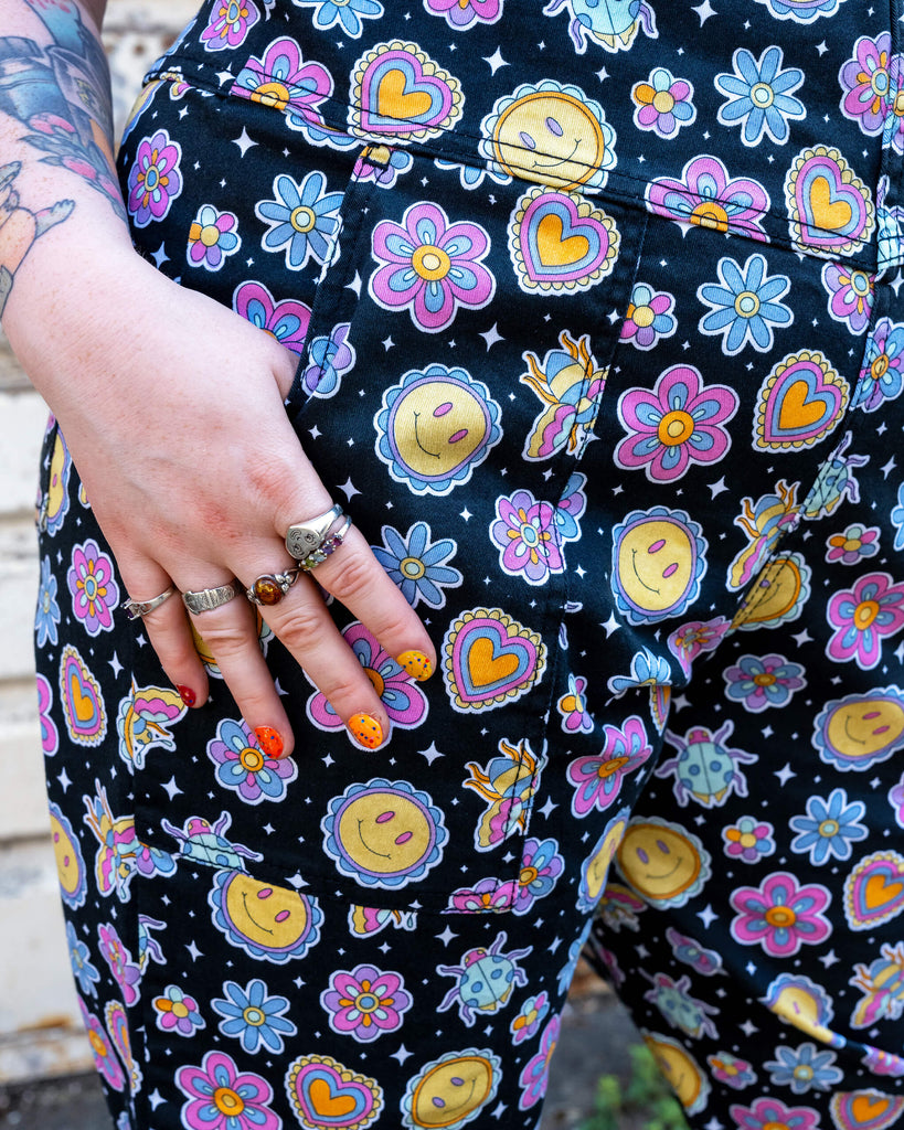 Close-up of a hand wearing silver rings resting on the pocket of a pair of black dungarees featuring a colourful, retro-inspired pattern of smiley faces, flowers, hearts, and stars. The person's nails are painted with bright, funky designs. The dungarees are from Run & Fly, a small ethical slow fashion brand known for their quirky, vibrant prints. The tattooed arm and cheerful outfit convey a playful, alternative style, reflecting the brand’s commitment to creativity and ethical production.