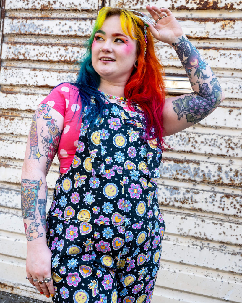 Person with rainbow hair and colourful makeup smiling in Run & Fly dungarees featuring a vibrant pattern of smiley faces, flowers, and hearts. Paired with a pink polka dot top, the outfit showcases a playful, alternative style. Run & Fly is a small ethical slow fashion brand known for its quirky, bold prints. The background is a rusted metal wall, enhancing the contrast between the lively fashion and the industrial setting.