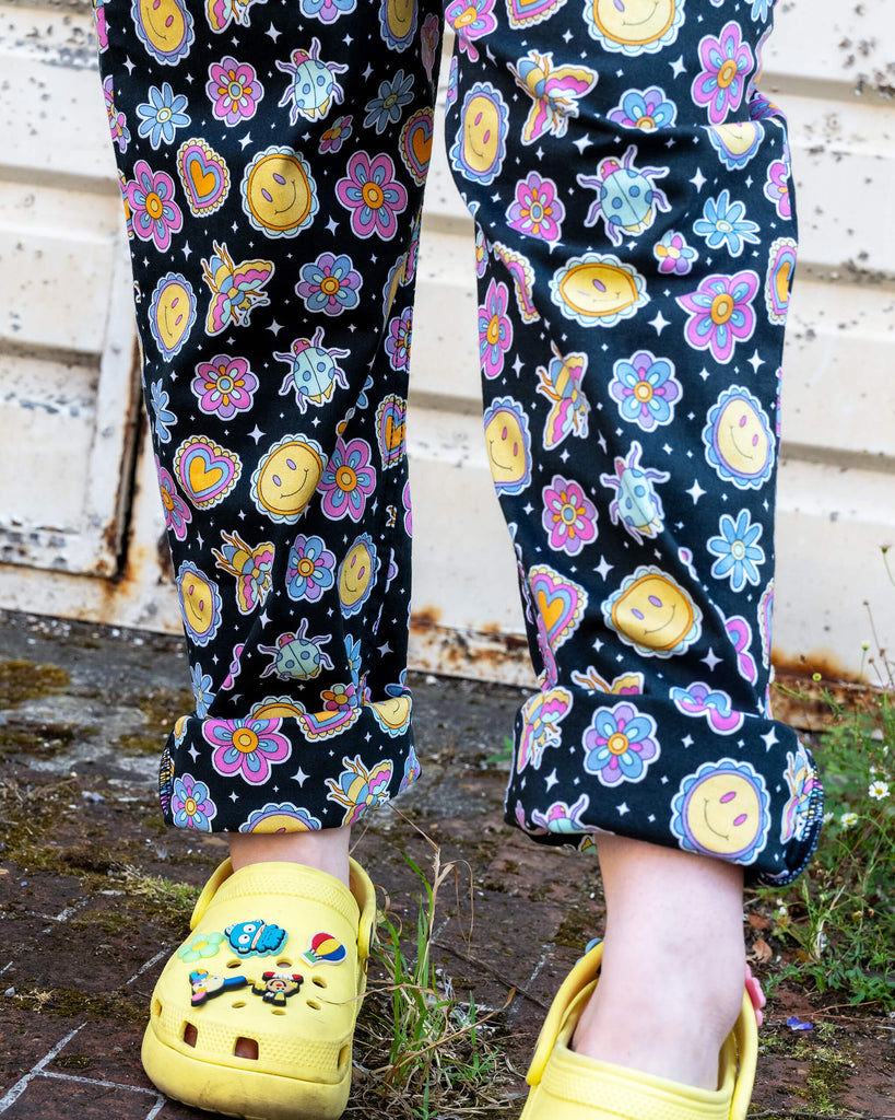 Close-up of legs wearing black Run & Fly dungarees with a retro smiley face and flower pattern. Paired with yellow Crocs decorated with playful charms, the outfit reflects a fun, alternative style. The ethical slow fashion brand is known for its quirky, colourful prints. The background features a textured, rusted metal wall, adding contrast to the vibrant look.