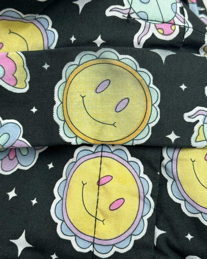 Close-up of a section of the Bee Happy Stretch Jumpsuit by Run & Fly, highlighting printing defects. A yellow smiley sun on the black fabric appears discoloured, with faded and uneven colouring.