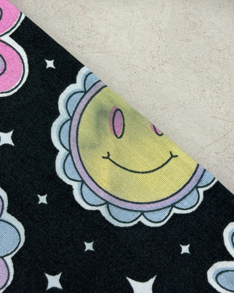 Close-up of the Bee Happy Stretch Jumpsuit fabric from Run & Fly, a small ethical slow fashion brand. The black fabric features a pastel smiley sun and star print, but there is a noticeable dark smudge on the yellow face, indicating a printing defect.