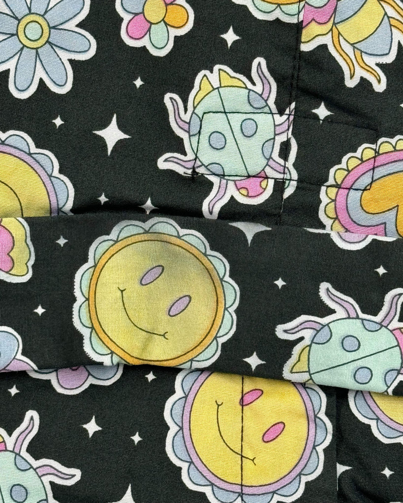 Fabric detail of Run & Fly’s Bee Happy Stretch Jumpsuit showing smiley suns, stars, and pastel bugs on a black background. The print has a fault, with an uneven yellow tone and dark patches on the sun design.