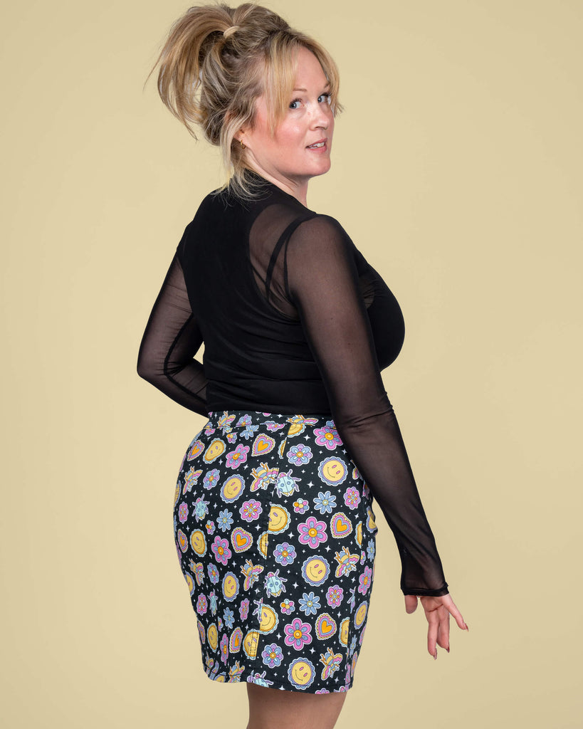 Karina showcases the playful Bee Happy mini skirt by Run & Fly, designed by Waffle Mama Design. The skirt features a lively pattern of smiley faces, hearts, and florals with a flattering A-line silhouette.