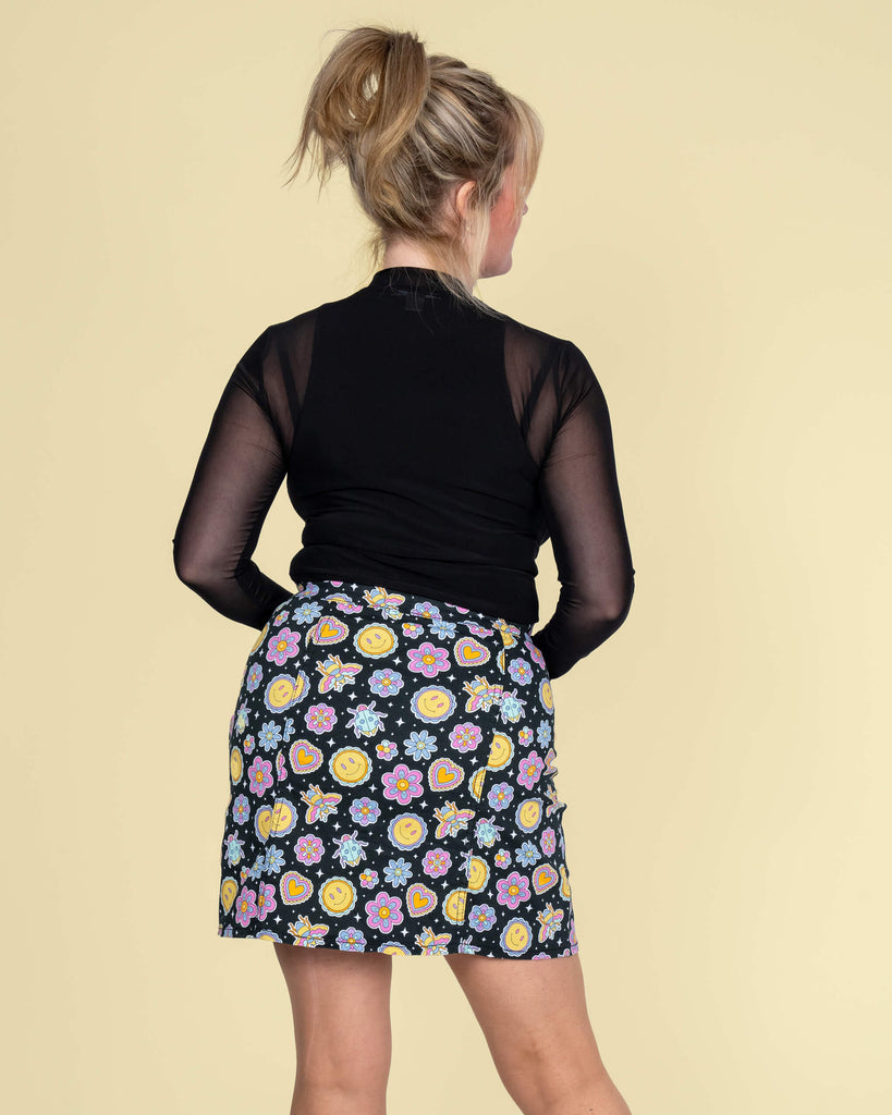  Back view of Karina wearing the Run & Fly mini skirt by Waffle Mama Design. The colourful print showcases smiley faces, flowers, and hearts in a quirky, cheerful design, complemented by a black mesh top.