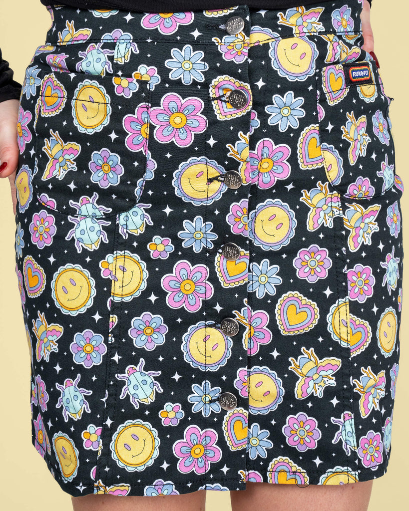 Close-up of the Run & Fly mini skirt print by Waffle Mama Design. The black fabric pops with bright illustrations of smiley faces, flowers, hearts, and buzzing bees, secured by branded buttons.