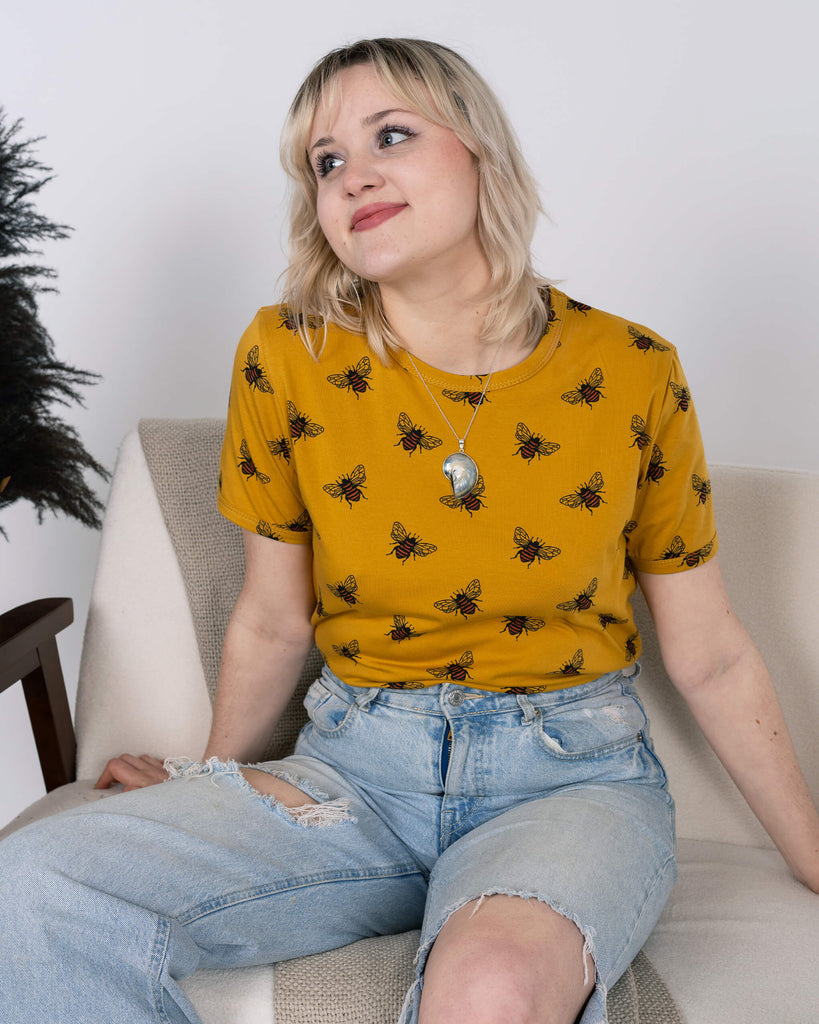 A person with blonde hair sits on a sofa, wearing the Run & Fly Buzzy Bee Mustard Gold Tee with an all-over bee print. Paired with ripped light-wash jeans, they lean slightly back, smiling softly. The mustard yellow t-shirt pops against the neutral tones of the room, emphasising its ethical slow fashion credentials.