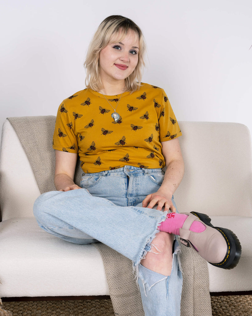 A person with blonde hair sits cross-legged on a cream-coloured sofa, wearing the Run & Fly Buzzy Bee Mustard Gold Tee. The t-shirt features an all-over bee print on mustard yellow fabric, styled with light-wash ripped jeans and pink socks paired with chunky shoes. The scene includes cosy textures and a neutral background, highlighting the sustainable and ethical design of the outfit.