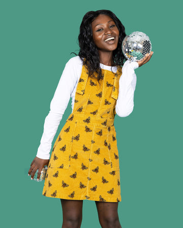 Close up of the Mustard Stretch Corduroy The Bees Kneez Bee Print Pinafore Dress worn by a femme alternative model with mid length black hair with a white long sleeve tshirt on a teal studio background. She is facing forward laughing to camera holding up two mini disco balls. The mustard gold yellow base pinafore features realistic repeating bees all over.