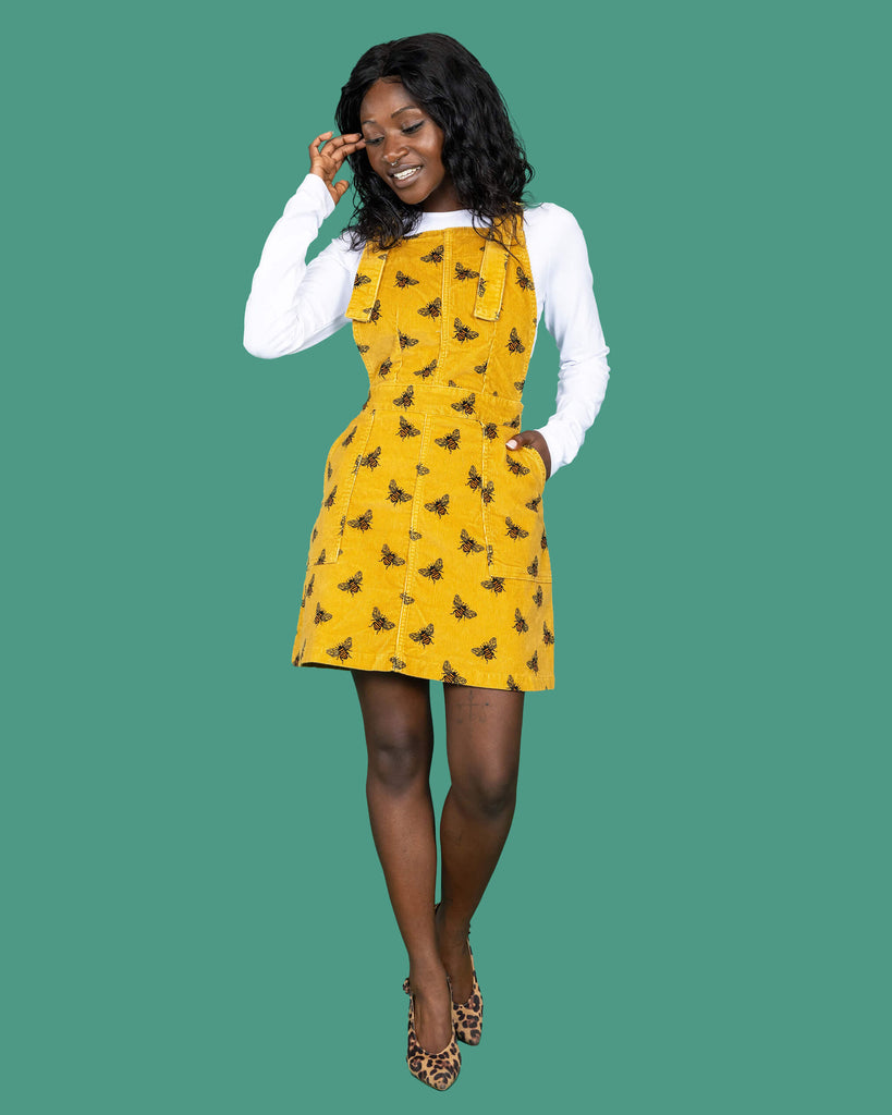 The Mustard Stretch Corduroy The Bees Kneez Bee Print Pinafore Dress worn by a femme alternative model with mid length black hair with a white long sleeve tshirt and leopard print heels on a teal studio background. She is facing forward laughing looking down to the left with one hand resting in the pinafore pocket and the other pushing back the side of her hair. The mustard gold yellow base pinafore features realistic repeating bees all over.
