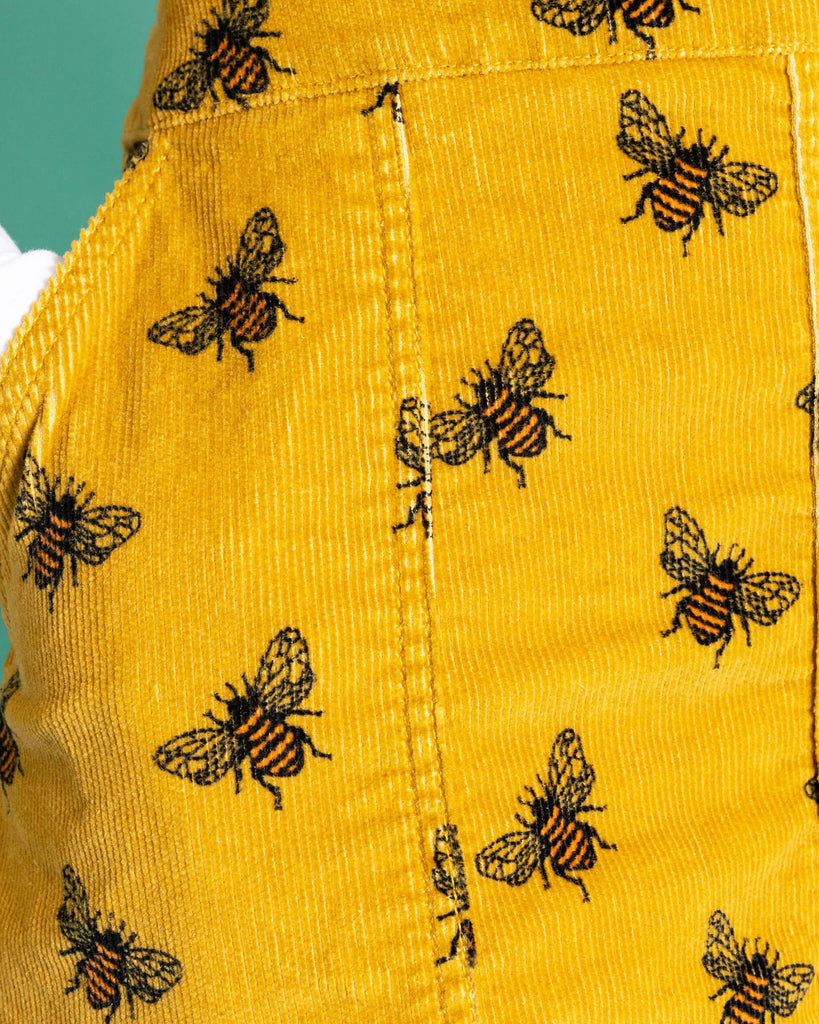 Close up of the Mustard Stretch Corduroy The Bees Kneez Bee Print Pinafore Dress worn by a femme alternative model with a white long sleeve tshirt on a teal studio background. She is facing forward with one hand resting in the pinafore pocket. The mustard gold yellow base pinafore features realistic repeating bees all over.