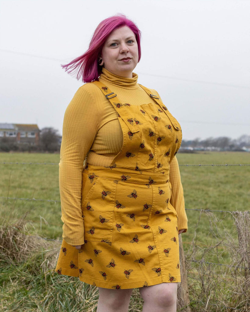 Emy-C is stood in front of a grass field wearing the bees knees bee print gold stretch twill pinafore dress with a long sleeve yellow tshirt and black cat boots. They are facing forward posing with both hands resting by their side.