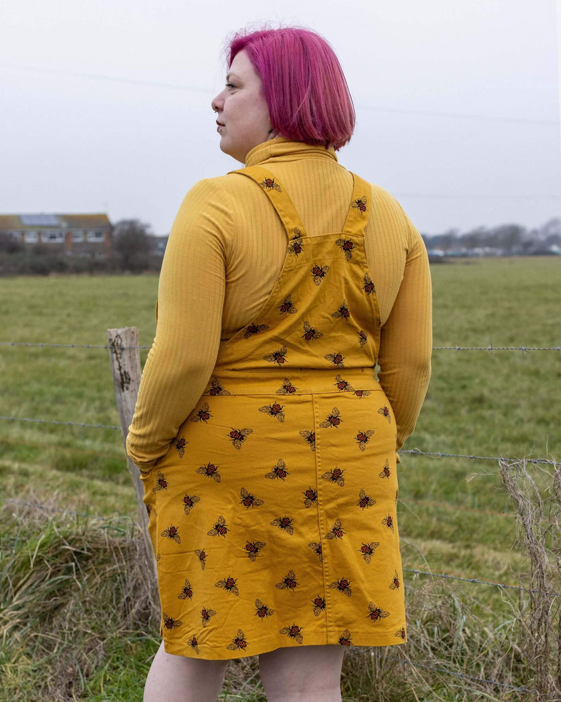 Emy-C is stood in front of a field area wearing the bees knees bee print gold stretch twill pinafore dress with a long sleeve yellow tshirt and black cat boots. They are facing away from the camera with both hands in the pinafore pockets.