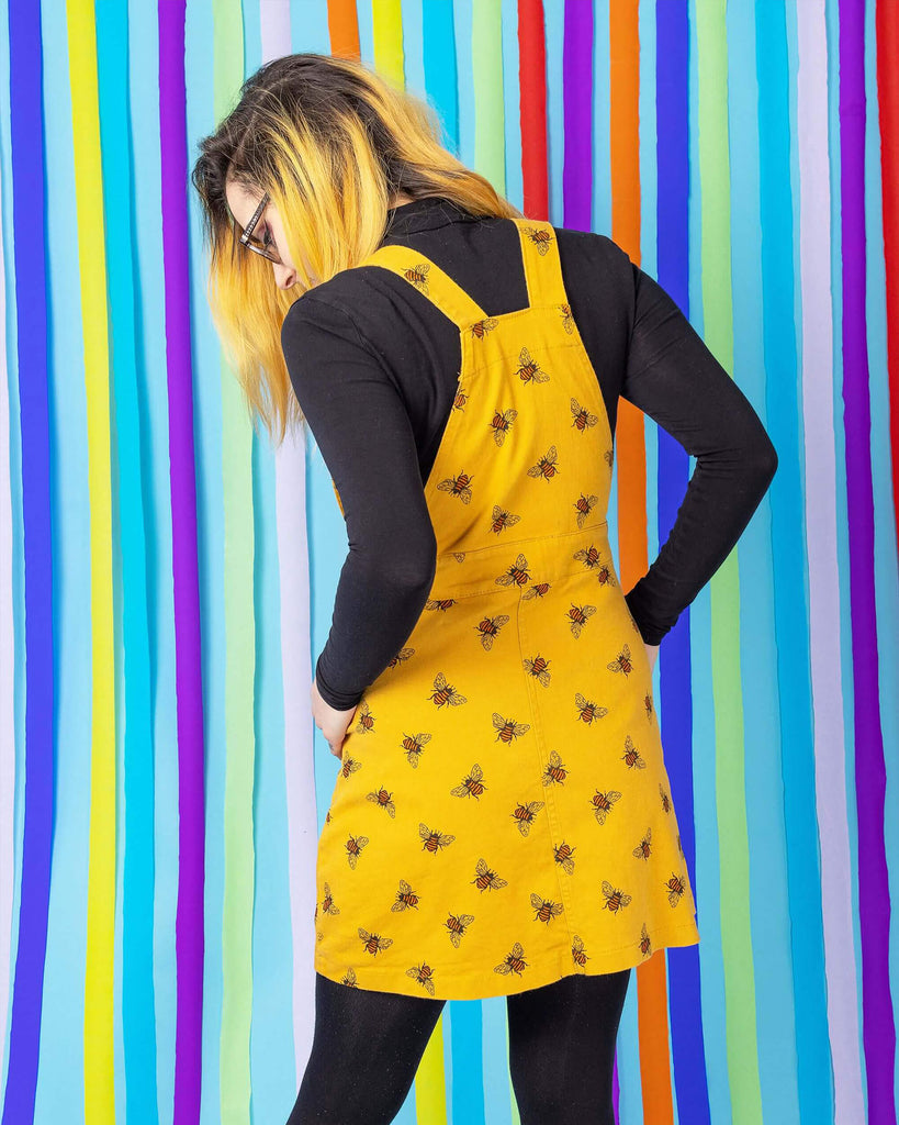 Jojo is stood in front of a blue rainbow studio background wearing the bees knees bee print gold stretch twill pinafore dress with a long sleeve black tshirt and grey boots. She is facing away from the camera with both hands in her pinafore pockets whilst looking down to the left.