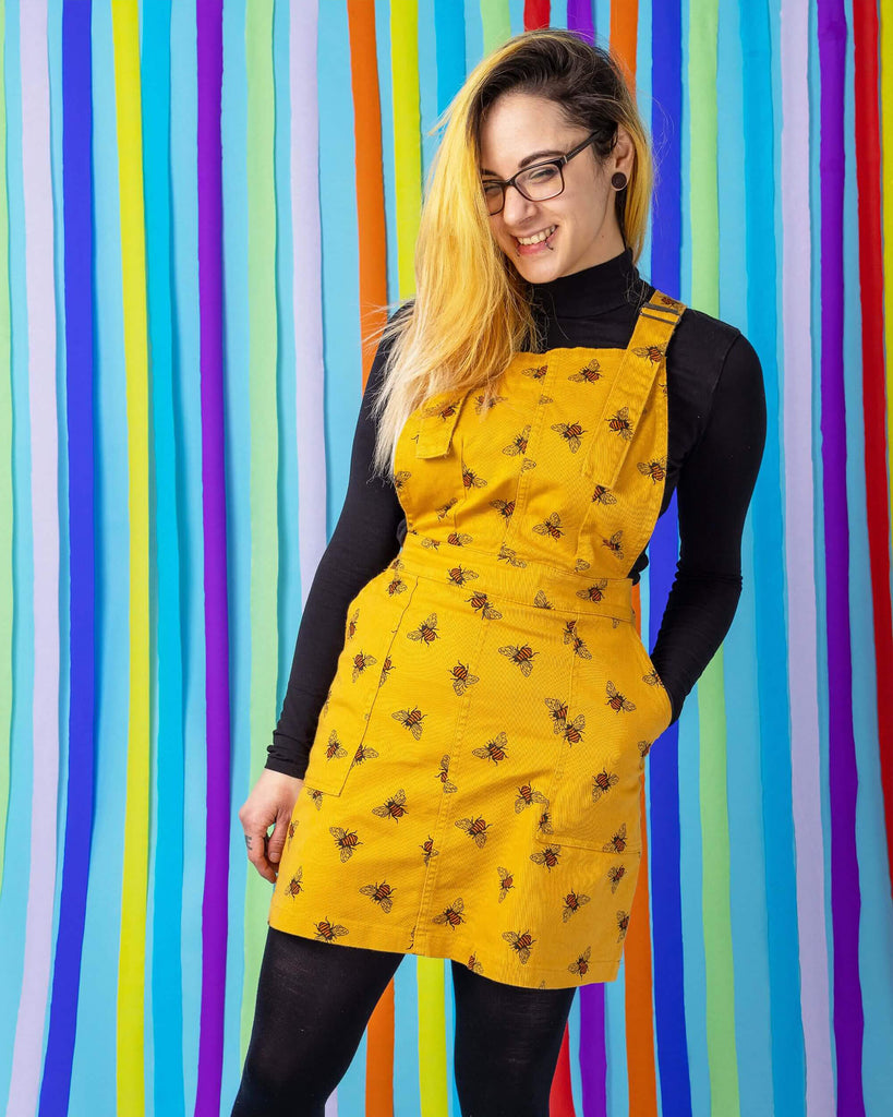 Jojo is stood in front of a blue rainbow studio background wearing the bees knees bee print gold stretch twill pinafore dress with a long sleeve black tshirt and grey boots. She is facing forward leaning back on one leg with one hand in her pinafore pocket whilst smiling to camera.