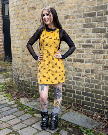 A model poses against a brick wall wearing Run & Fly’s Gold Bee Corduroy Pinafore Dress, a mustard yellow non-stretch corduroy design with an embroidered black and orange bee print. The fitted waist and adjustable buckle straps create a flattering silhouette. Styled with a black mesh top, chunky black platform boots, and tattooed legs on show.