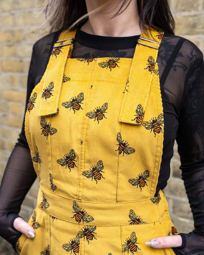 A detailed close-up of the Gold Bee Corduroy Pinafore Dress, showcasing the adjustable buckle straps and bold bee embroidery. The model wears a black mesh top underneath, adding an edgy contrast to the vintage-inspired design. Made from non-stretch corduroy for a structured fit.