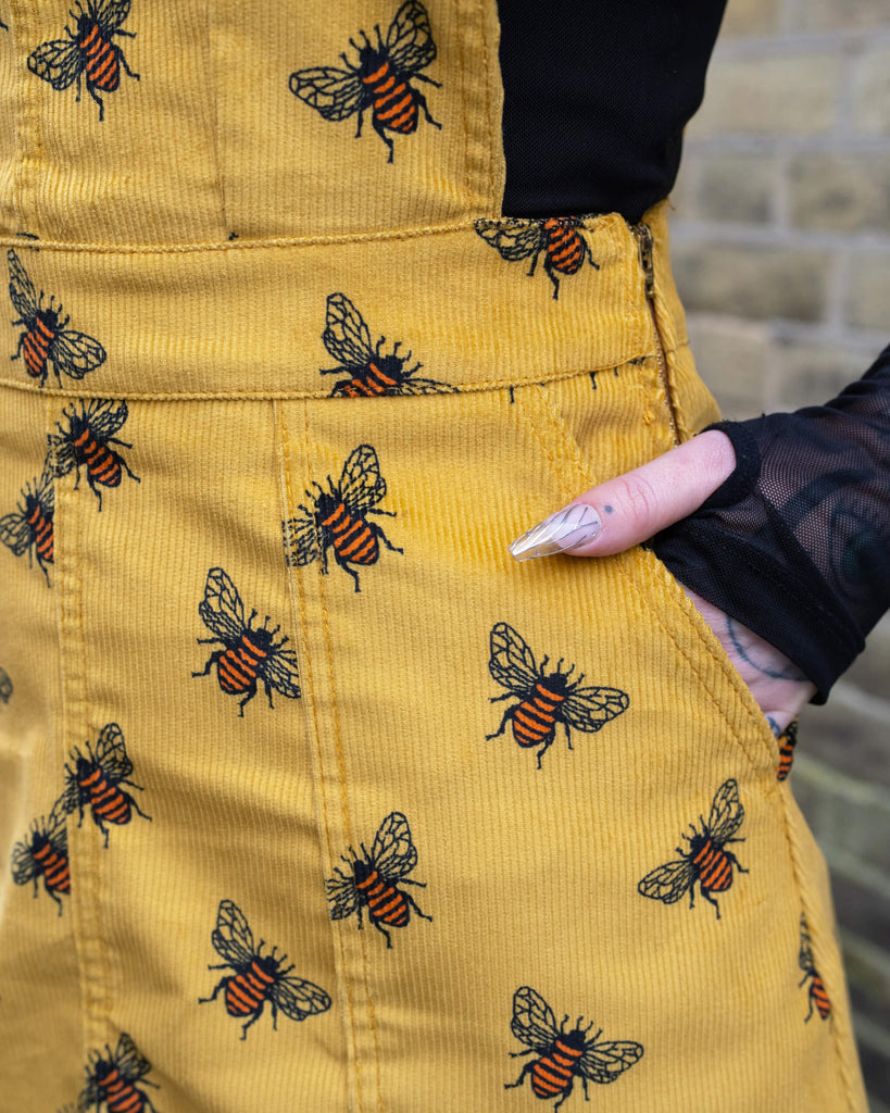 A close-up of the Gold Bee Corduroy Pinafore Dress by Run & Fly, highlighting the detailed embroidered bee print on mustard yellow non-stretch corduroy. The model places a hand in the deep side pocket, showing off intricate nail art. Ethical slow fashion with a quirky twist.