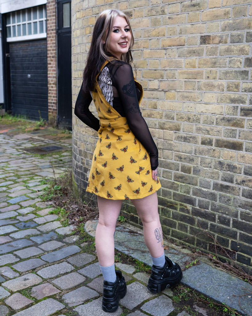 Back view of Run & Fly’s Gold Bee Corduroy Pinafore Dress, featuring a playful mustard yellow corduroy fabric with a bee print. The model poses on a cobbled street, showing the adjustable cross-back straps and fitted waist detail. Styled with a black mesh top and alternative platform boots.