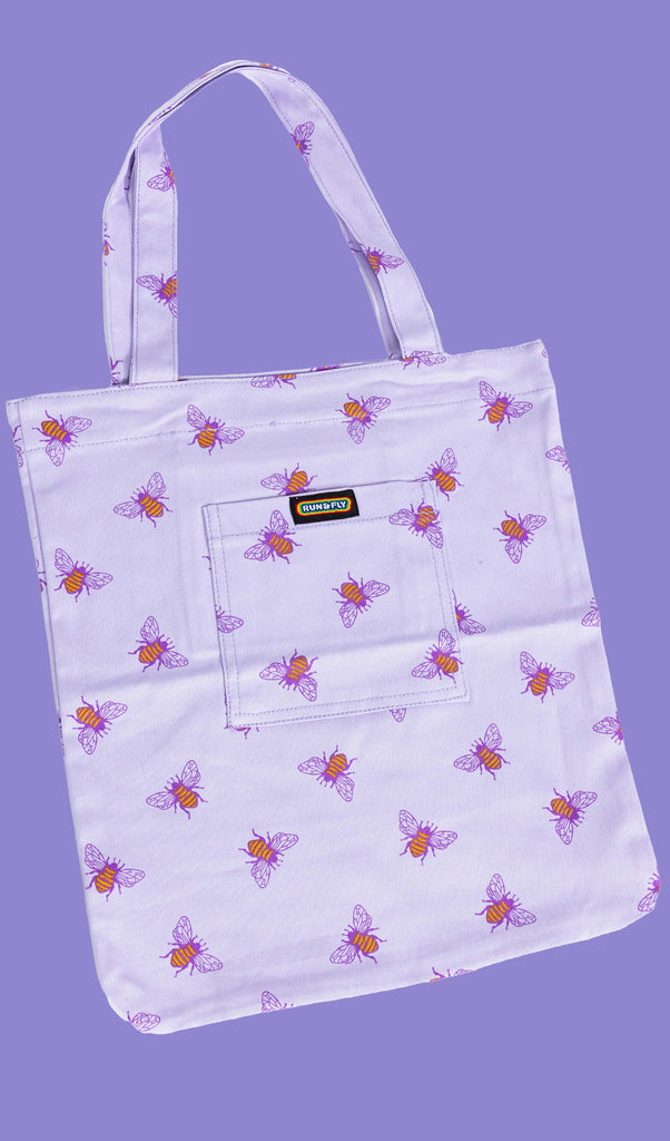 flat lay of a lilac tote bag with an all over print of yellow bumble bees with purple wings on a purple backdrop. There is a front pocket on the tote bag with Run&Fly logo on the front.