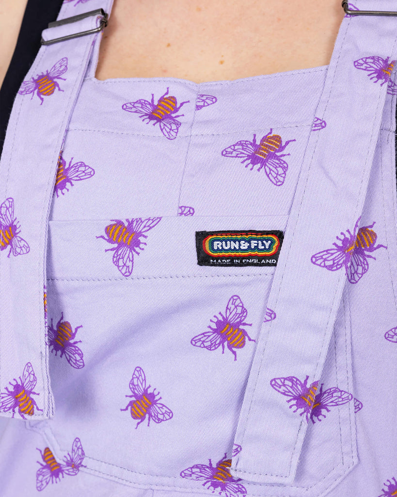 Close up of the Lavender Bees Stretch Twill Dungaree Shorts worn by a femme model with short blonde hair with a black crop top. She is facing forward to highlight the retro roller straps and the front bib pocket with a rainbow run & fly tag sewn in the top right corner. The dungaree shorts print is a pastel purple covered with dark purple and mustard yellow bee illustrations all over.