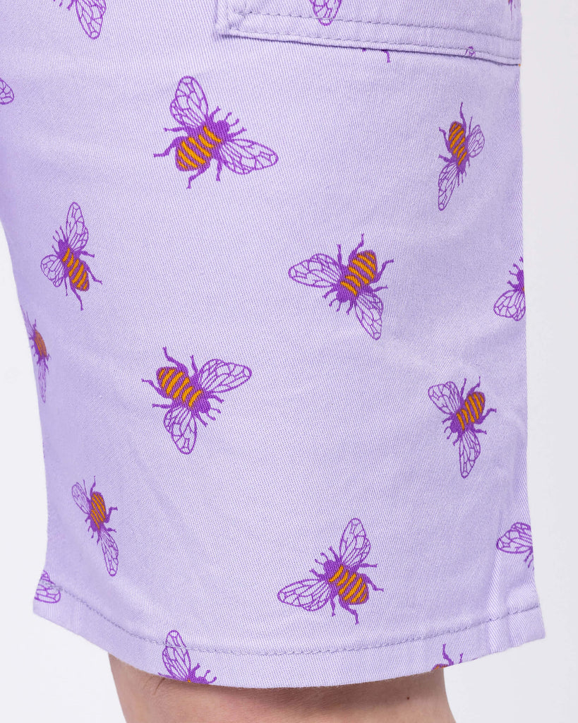Close up of the Lavender Bees Stretch Twill Dungaree Shorts worn by a femme model. She is facing forward to highlight the side pocket and short hem. The dungaree shorts print is a pastel purple covered with dark purple and mustard yellow bee illustrations all over.