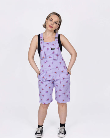 The Lavender Bees Stretch Twill Dungaree Shorts worn by a femme model with short blonde hair with a black crop top and black trainers. She is facing forward smiling with her head tilted slightly, both hands are resting on her hips in the dunga short pockets. The dungaree shorts print is a pastel purple covered with dark purple and mustard yellow bee illustrations all over.