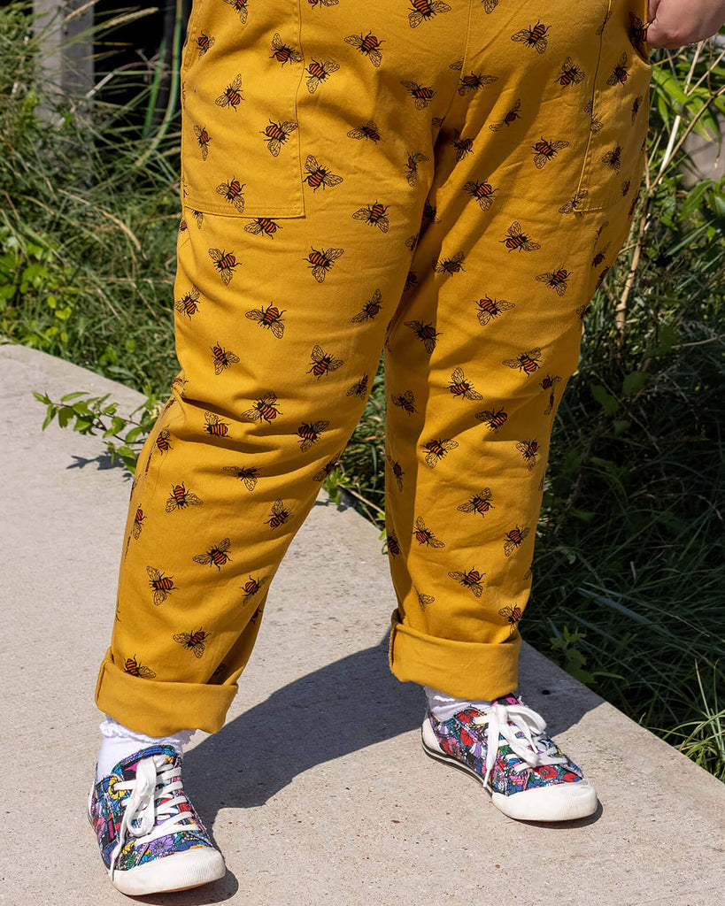 Close up of Natalie's legs wearing the dungabee gold stretch twill dungarees with multicoloured trainers. She is stood outside facing forward on a concrete path in shoreham.