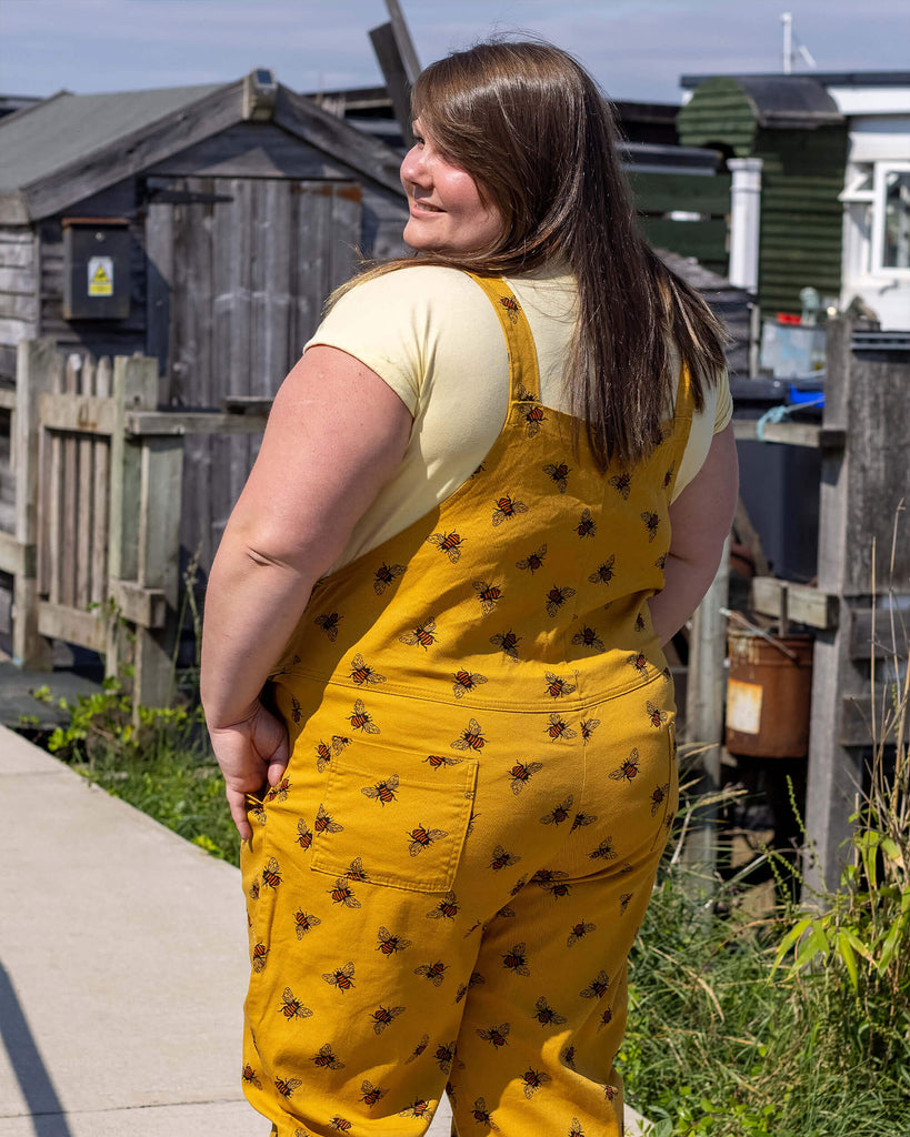 The back of Natalie wearing the Bees Knees Gold Stretch Twill Dungarees with a yellow tee and canvas trainers. She is facing away from the camera with her hand in her pocket.