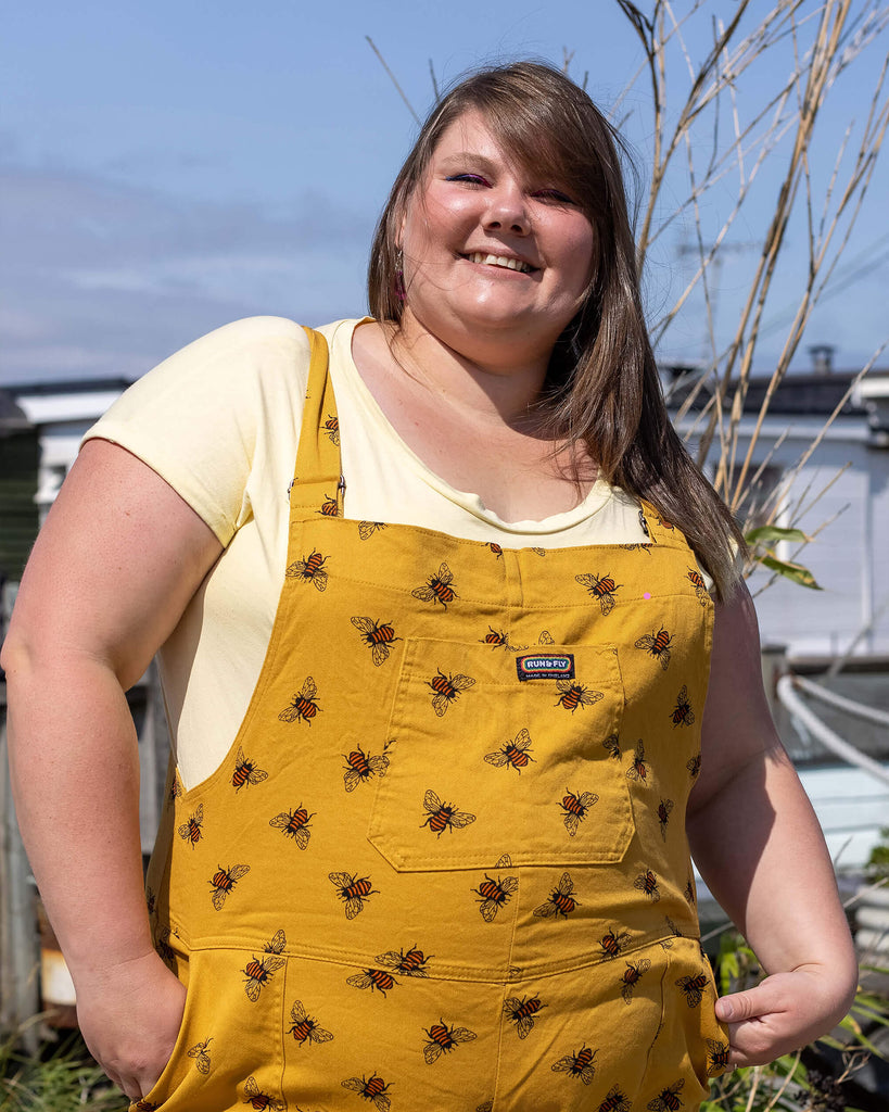 Natalie wearing the Bees Knees Gold Stretch Twill Dungarees with a yellow tee. She is smiling at the camera with her hands in her pockets.
