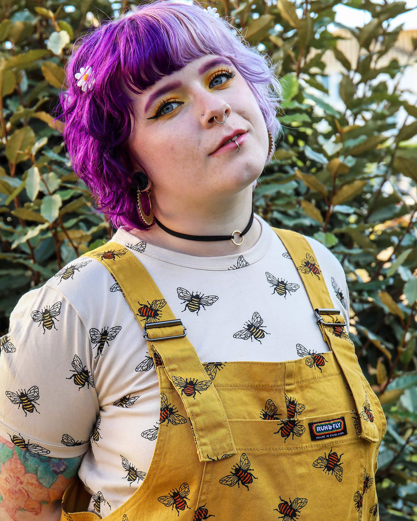 Georgia a purple haired white model with yellow eye make up and cute jewellery is wearing the Bees Knees Gold Stretch Twill Dungarees with the buzzy bee tee.