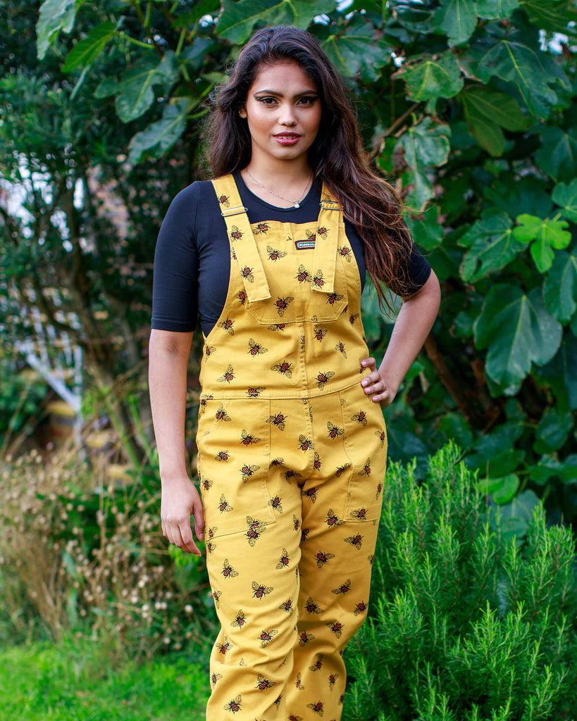 Jarin wearing the Bees Knees Gold Stretch Twill Dungarees with a black tee and platform black boots. She has her hand on her hip and is looking into the camera.