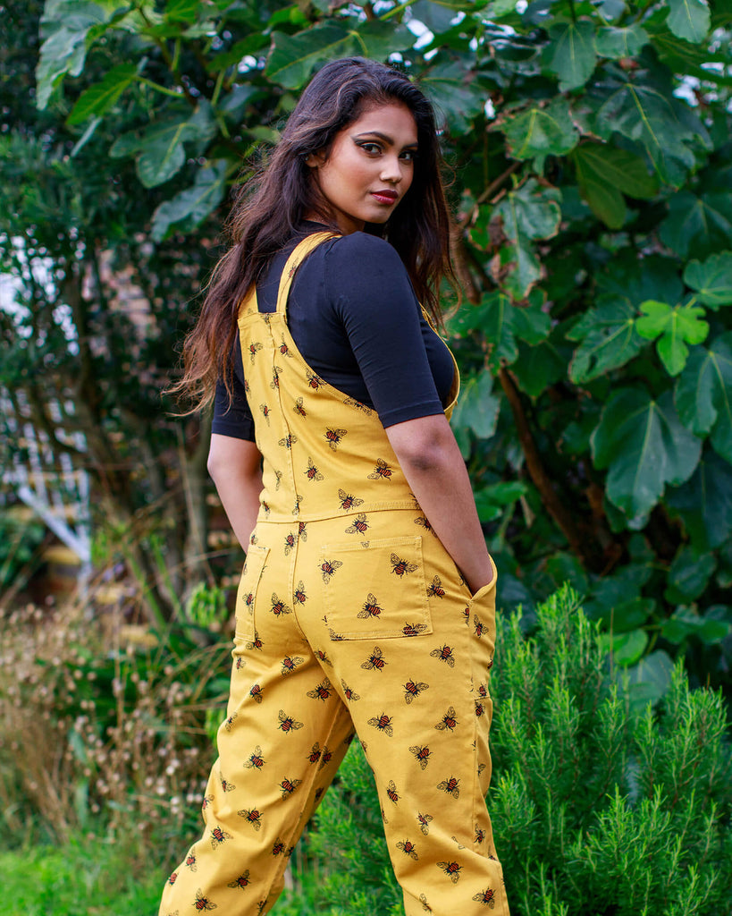 The back of Jarin wearing the Bees Knees Gold Stretch Twill Dungarees with a black tee and platform black boots. She has her hands in her pockets and she is looking over her shoulder.