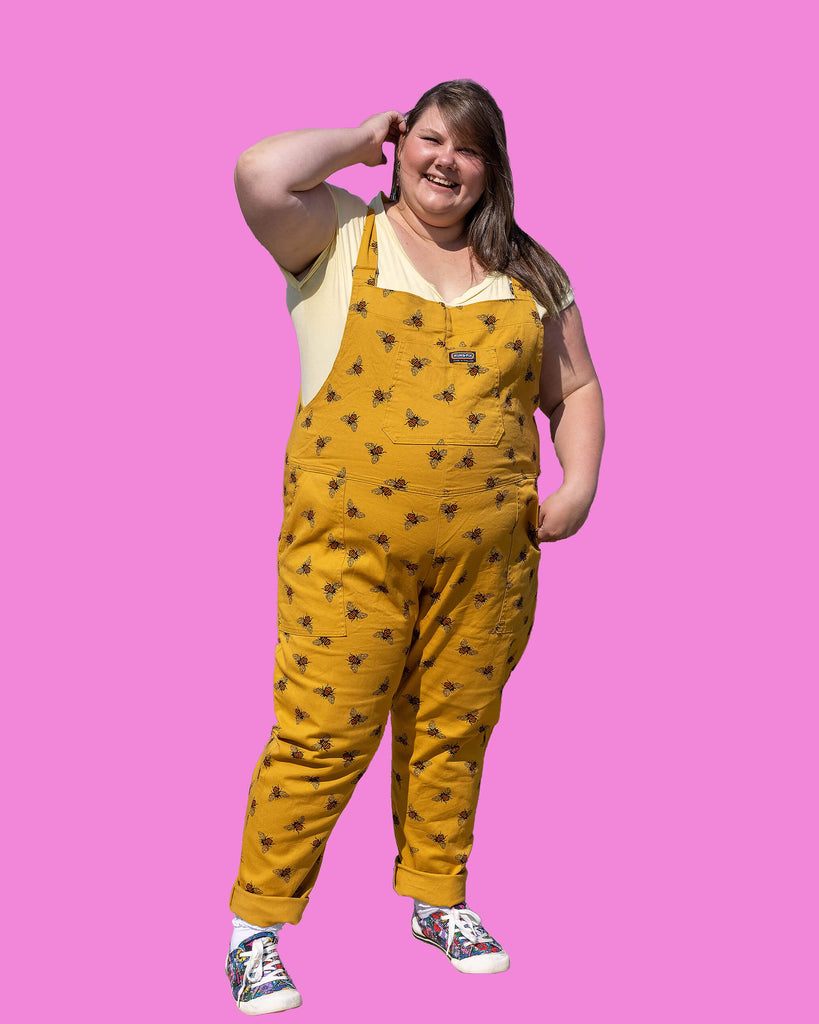 Full length photo of Natalie on a pink background wearing the Bees Knees Gold Stretch Twill Dungarees with a yellow tee and canvas trainers. Her hand is pushing her hair out of her face and she's smiling at the camera.