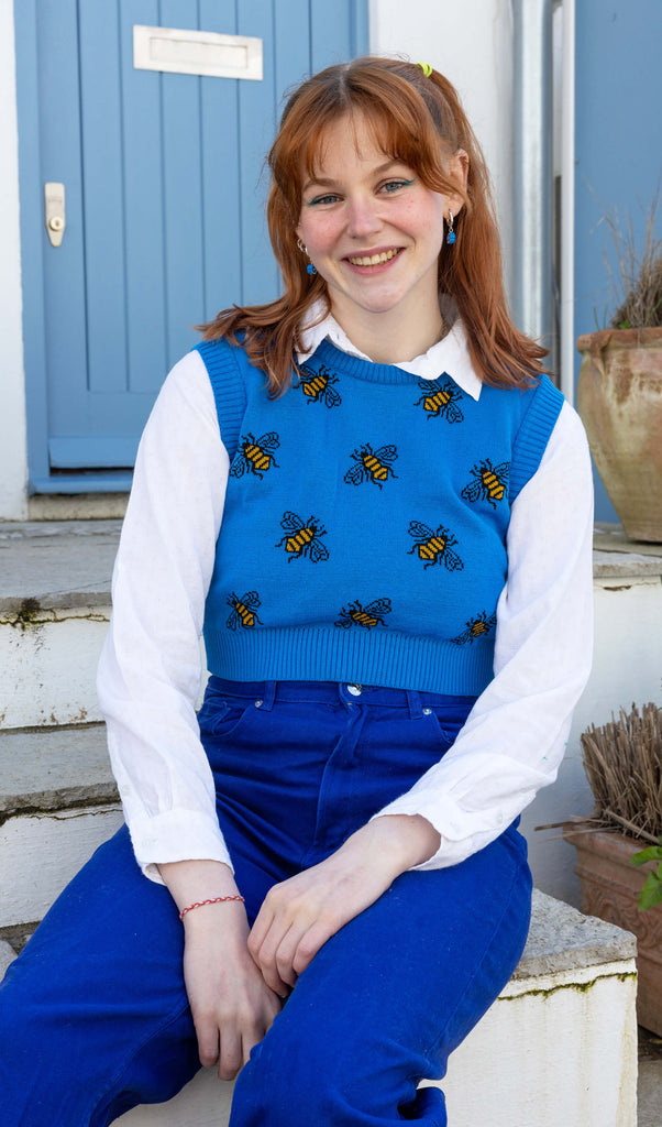 Tasha a femme red head with short hair and a fringe is smiling at camera wearing a blue cropped bee knitted top with a white shirt and bright blue jeans. She is sat by a blue door