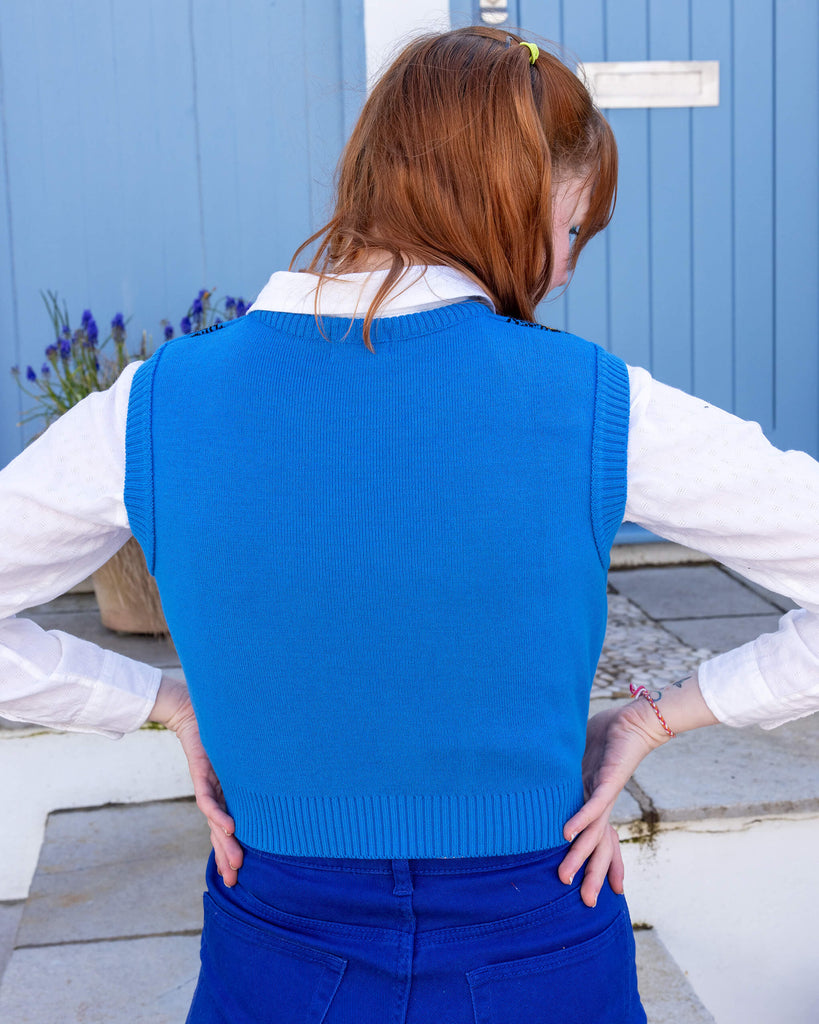 Tasha a femme red head with short hair and a fringe is smiling at camera wearing a blue cropped bee knitted top with a white shirt and bright blue jeans. She is facing the back with her hands on her hips