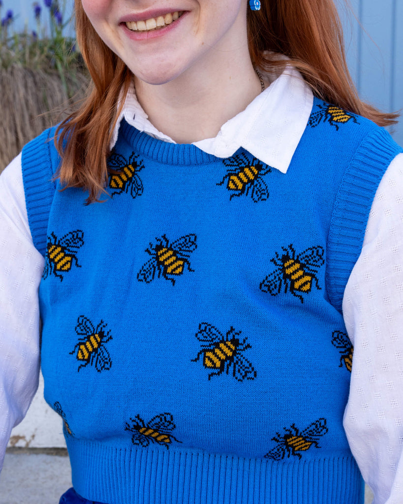 Tasha a femme red head with short hair and a fringe is smiling at camera wearing a blue cropped bee knitted top with a white shirt and bright blue jeans. She is sat by a blue door